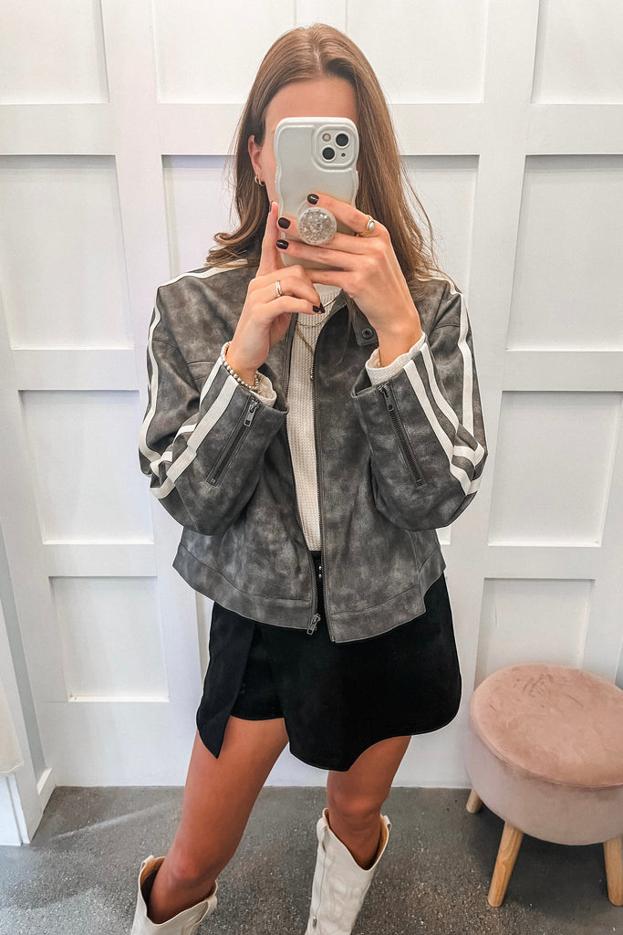 Person taking a mirror selfie wearing a gray jacket with white stripes, black shorts, and white boots.