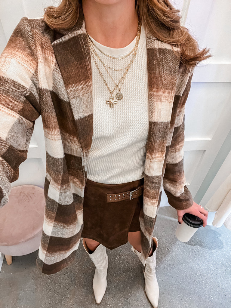 Person wearing a plaid cardigan, white sweater, brown shorts, and white boots, holding a coffee cup.