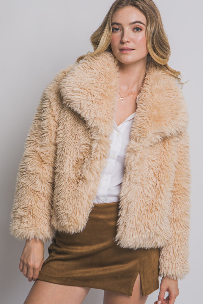 Woman wearing a beige fluffy jacket over a white top and brown skirt on a plain background