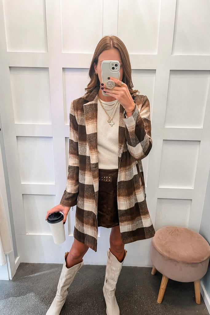 Person wearing a plaid coat, white sweater, and brown skirt with white boots, taking a mirror selfie.