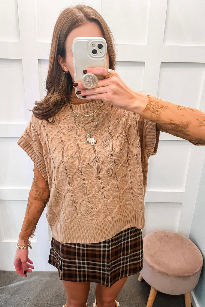 Woman taking a mirror selfie wearing a beige cable knit sweater vest and plaid skirt.