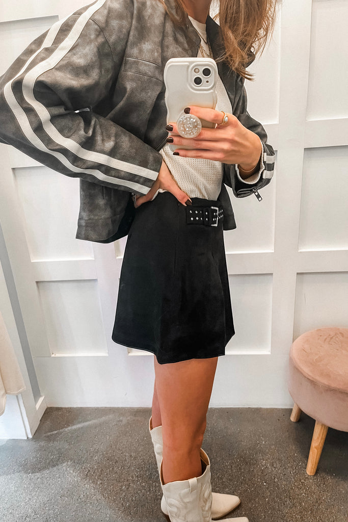 Person taking a mirror selfie wearing a gray jacket, black shorts, and white boots in a room with a white wall and pink stool.