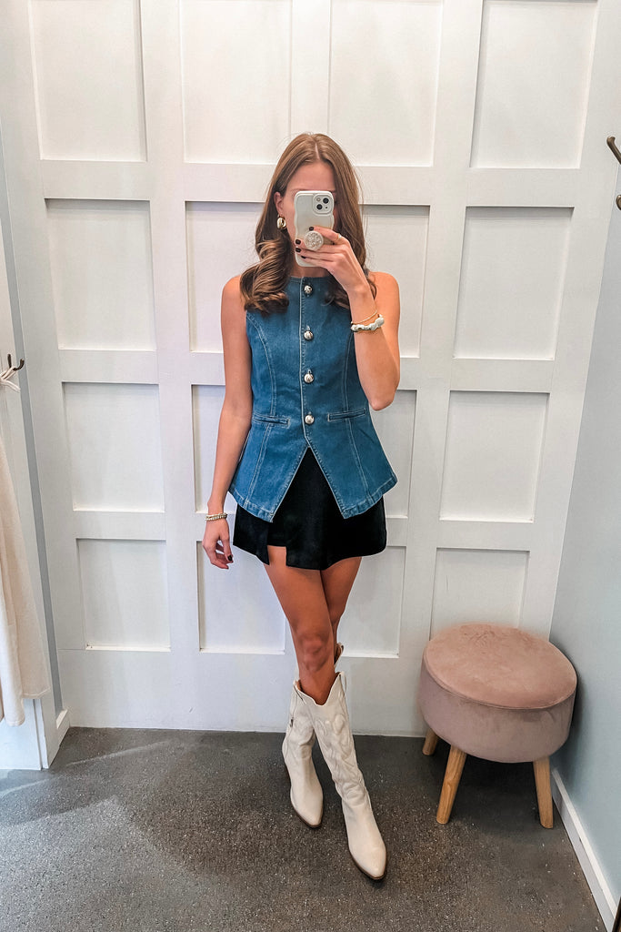 Person wearing a denim vest, black skirt, and white boots in front of a white paneled wall.