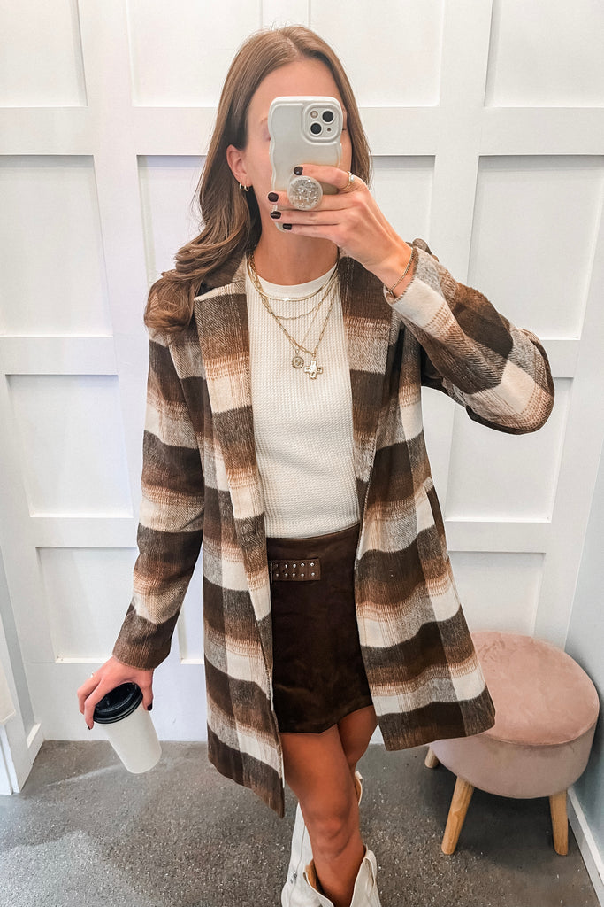 Woman taking a mirror selfie wearing a plaid coat, white sweater, and brown skirt.