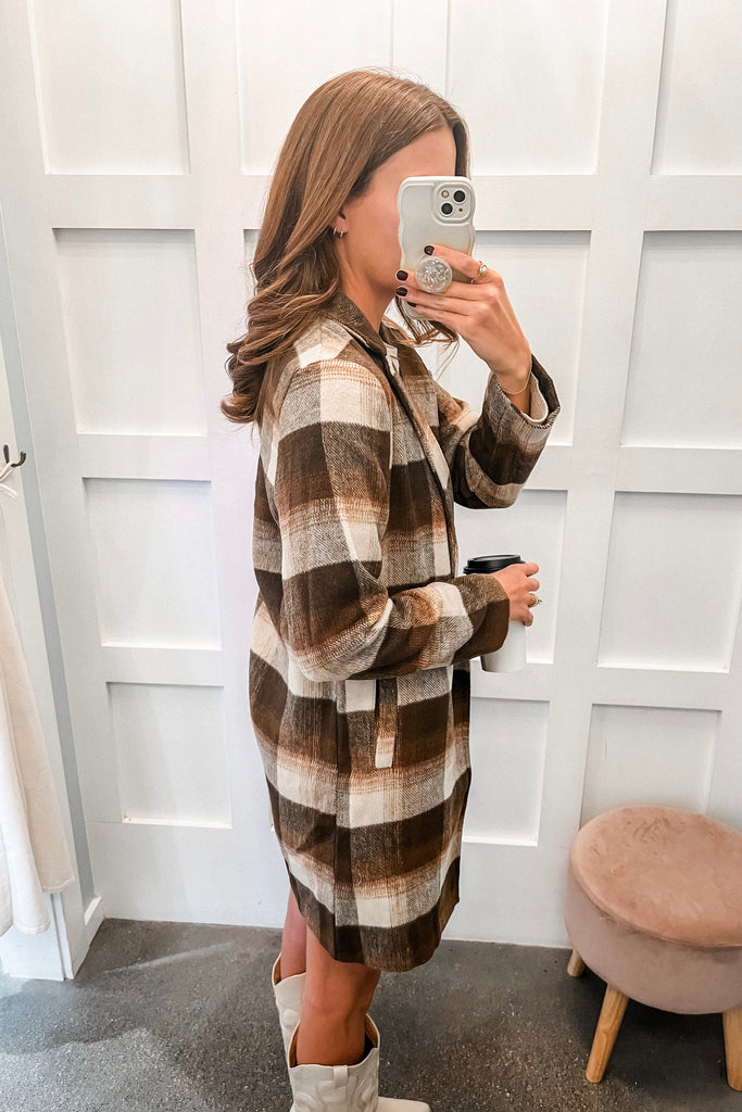 Woman wearing a plaid dress and white boots, taking a mirror selfie.