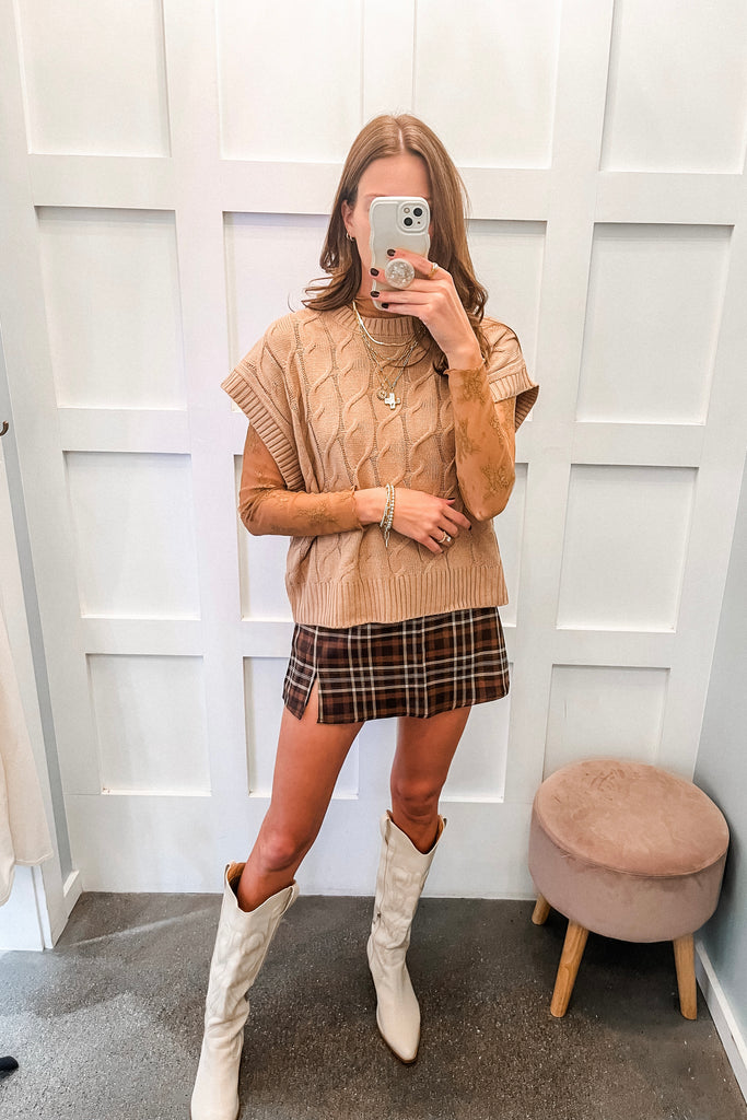 Woman taking a mirror selfie wearing a beige sweater, plaid skirt, and white knee-high boots in a room with white paneled walls.