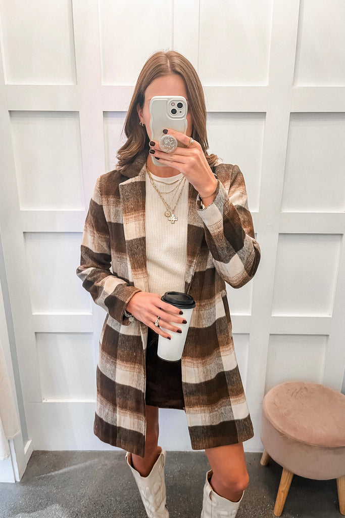 Person wearing a plaid coat, white boots, and holding a coffee cup in front of a white paneled wall.