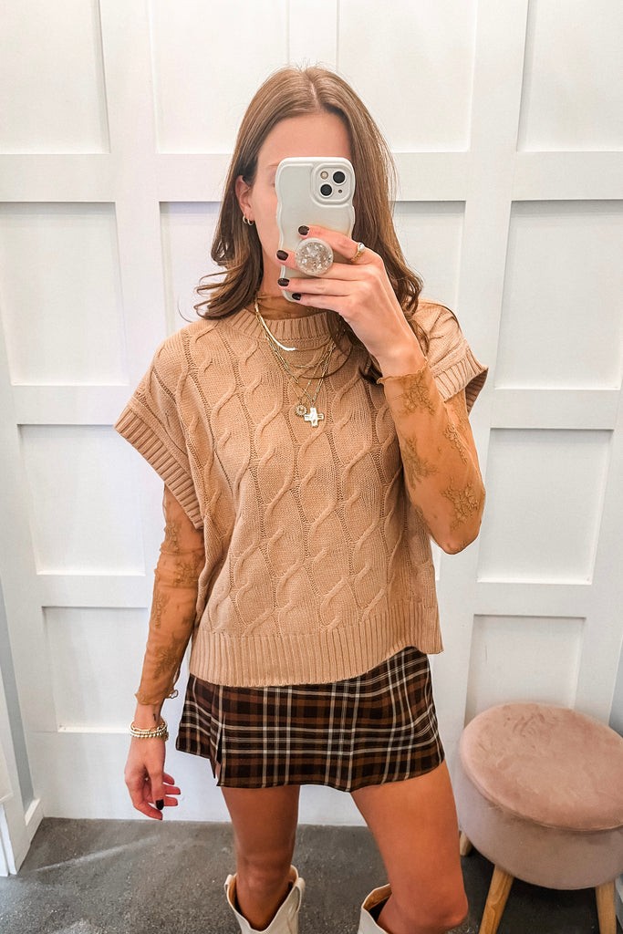 Woman taking a mirror selfie wearing a beige cable knit sweater and plaid skirt.