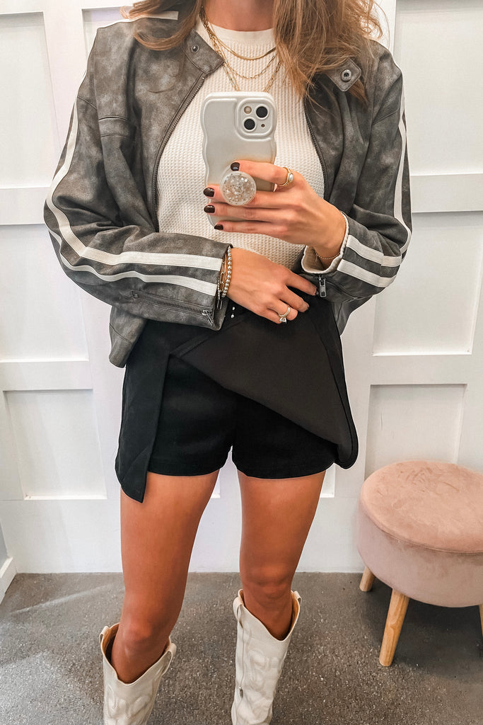 Person wearing a gray jacket with white stripes, white sweater, black shorts, and white boots, taking a mirror selfie.
