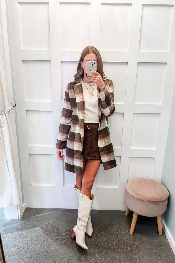 Person wearing a plaid coat, white shirt, brown skirt, and white boots in front of a white door.