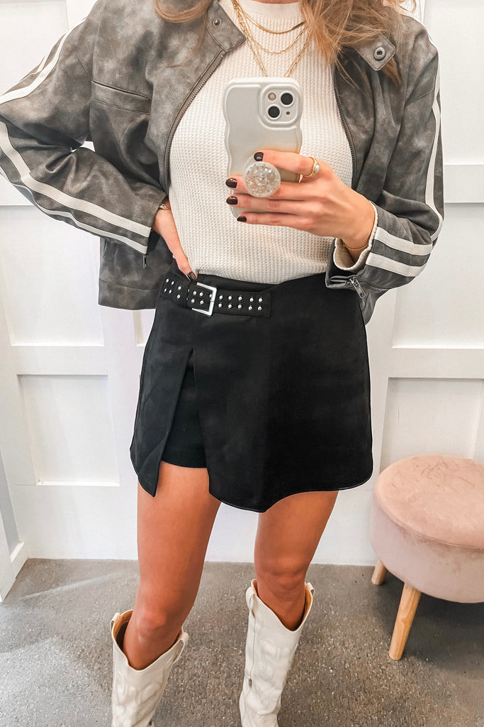 Person wearing a gray jacket with white stripes, white sweater, black skirt, and white boots, holding a phone.