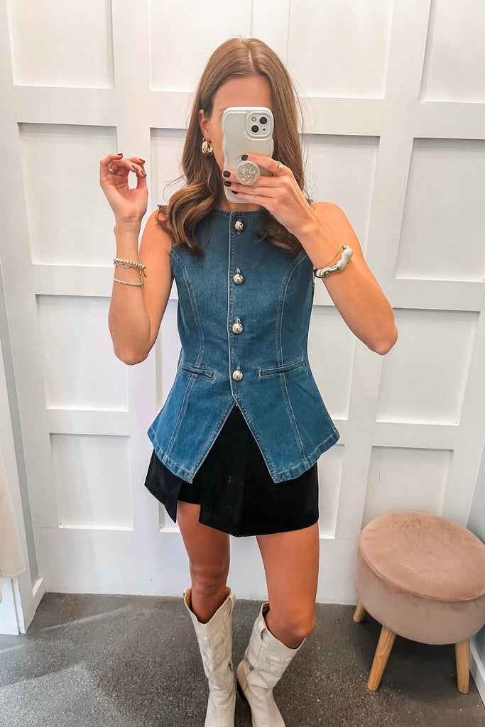 Woman taking a mirror selfie wearing a denim vest over a black dress with white boots.