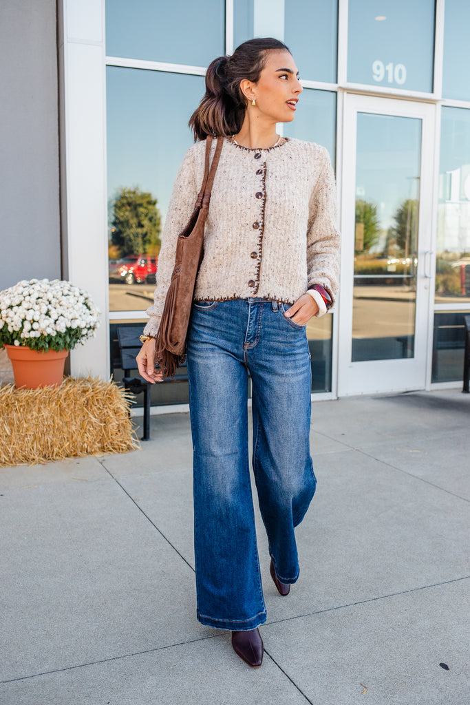 Woman in a beige cardigan and blue jeans walking outside a building.