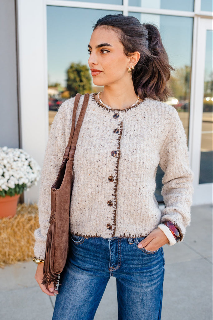 Woman wearing a beige cardigan and blue jeans, standing outdoors.