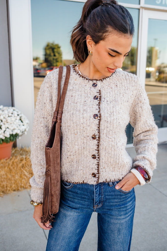 Woman wearing a textured beige cardigan with brown buttons, blue jeans, and carrying a brown bag.