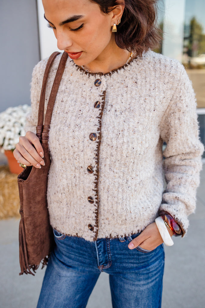 Woman wearing a textured beige cardigan with brown buttons, holding a brown bag.