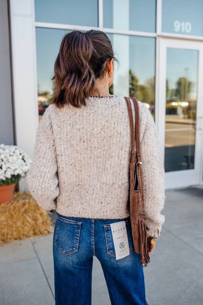 Person wearing a beige sweater and blue jeans with a brown bag, standing outside a building.