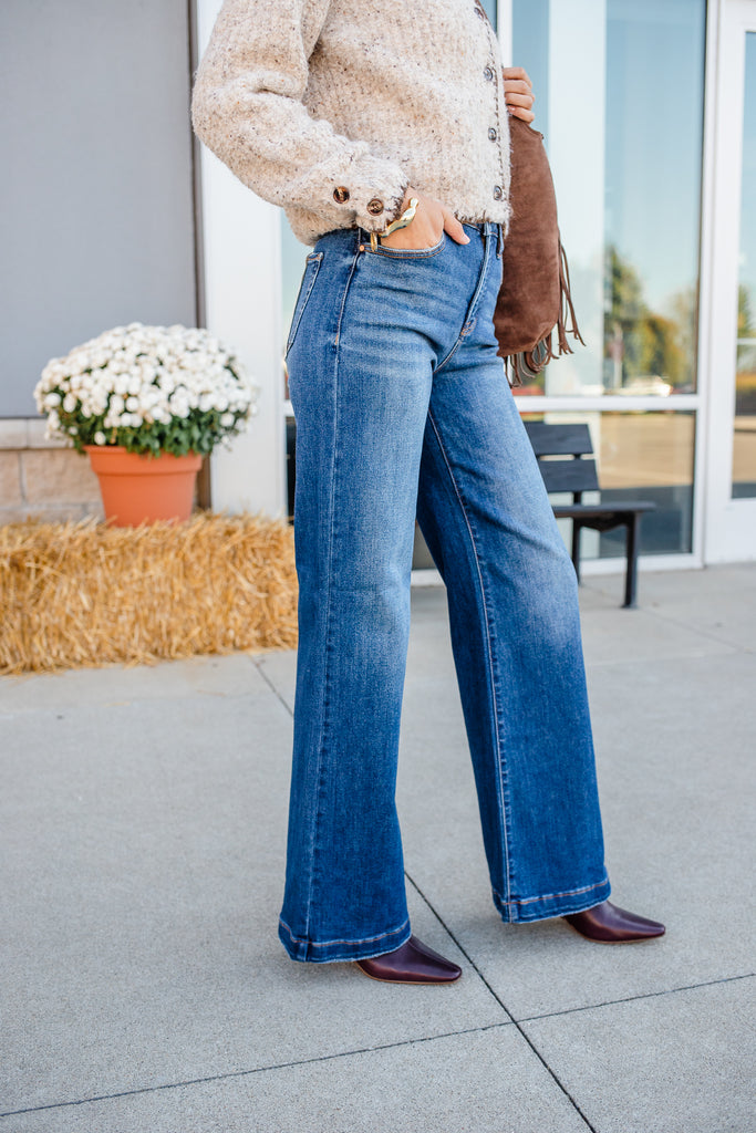 Person wearing blue jeans standing on a sidewalk with a building and flowers in the background