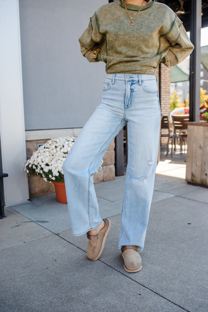 Person wearing a green sweater and light blue jeans standing on a sidewalk.