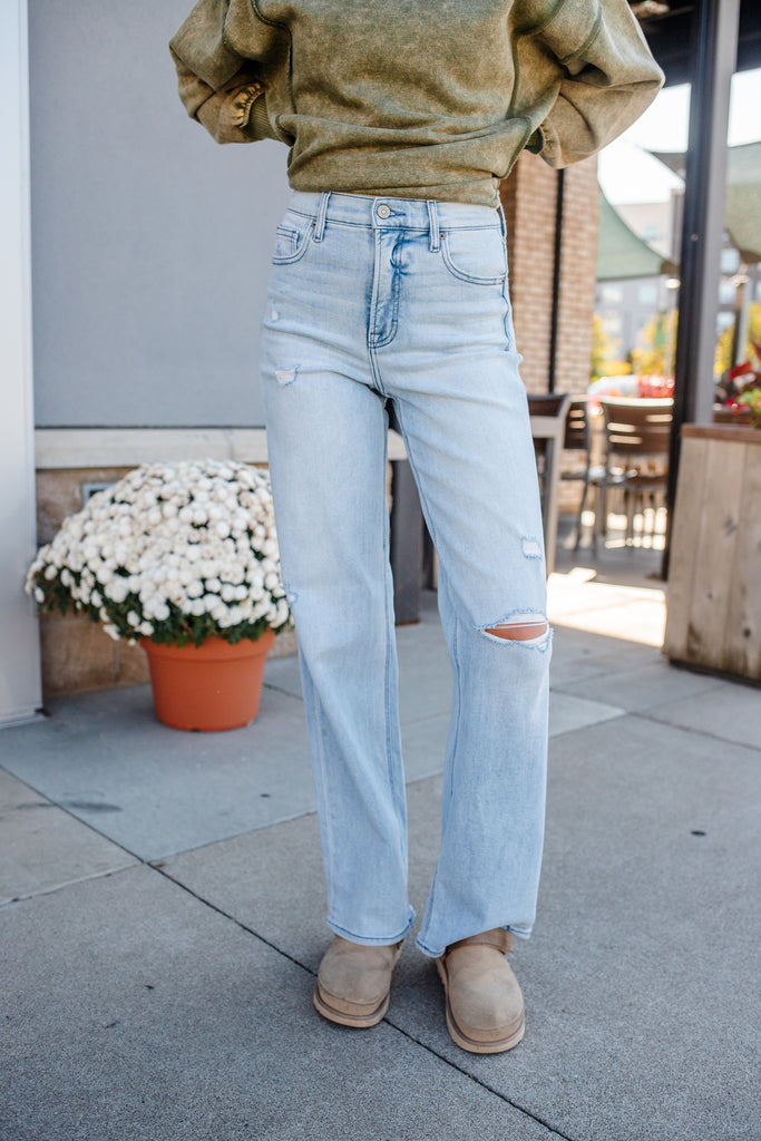 Person wearing light blue jeans and a green sweater standing on a sidewalk with a potted plant and building in the background.