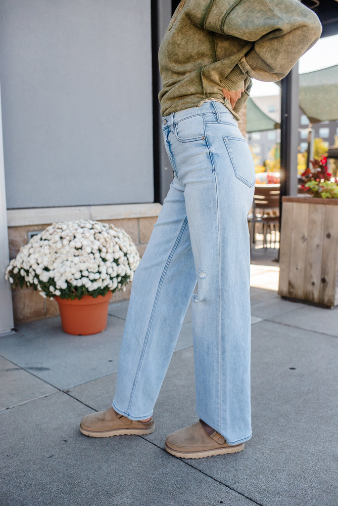 Person wearing light blue jeans and a green jacket outdoors with potted plants in the background