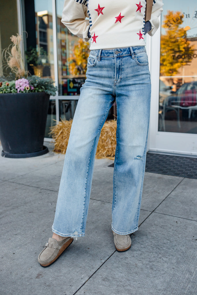 Person wearing a white sweater with red stars and blue jeans on a sidewalk.