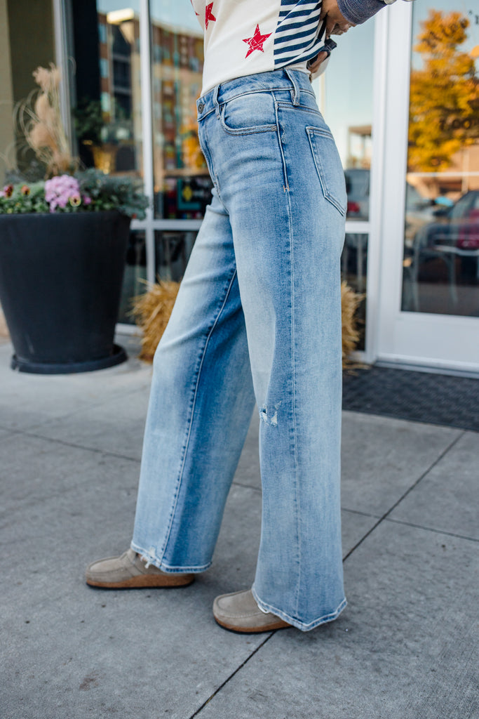 Person wearing light blue jeans standing on a sidewalk with a store in the background