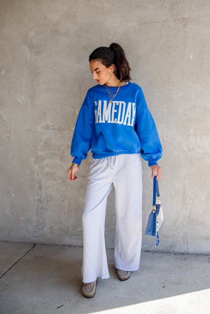 Woman wearing a blue 'GAMEDAY' sweatshirt and white pants against a concrete wall.