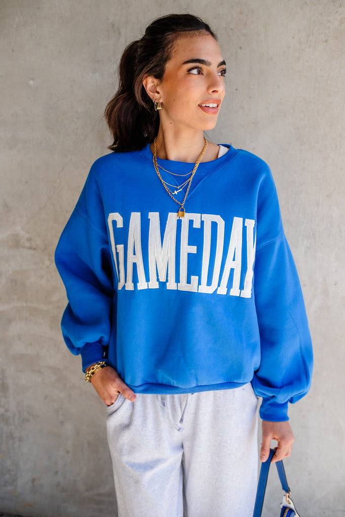 Woman wearing a blue 'GAMEDAY' sweatshirt against a gray wall