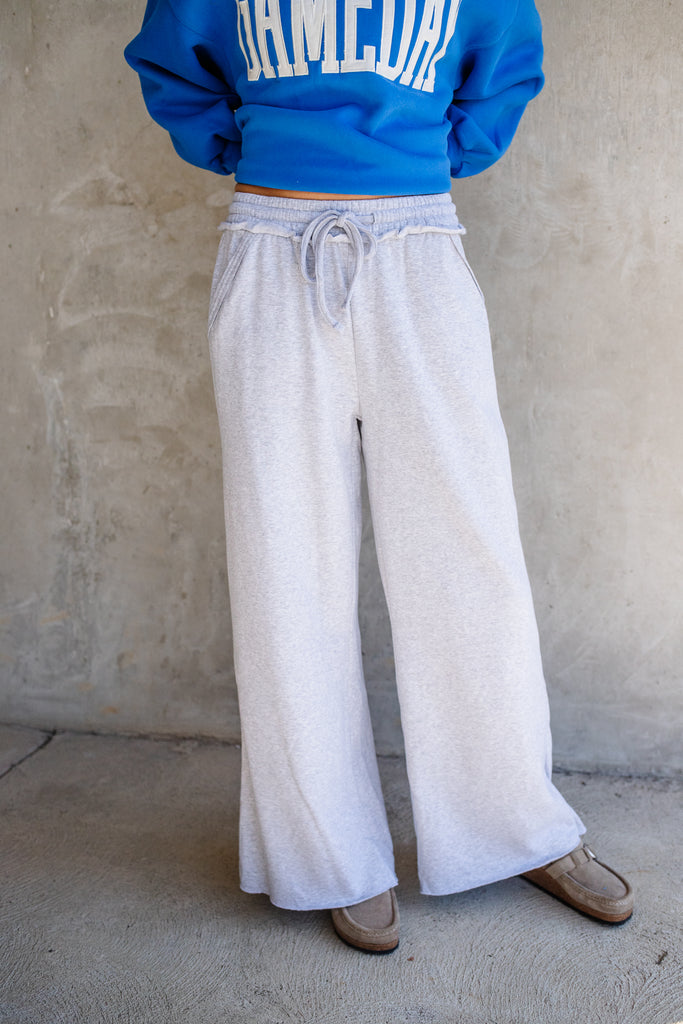 Person wearing a blue sweatshirt and light gray wide-leg pants against a beige wall.