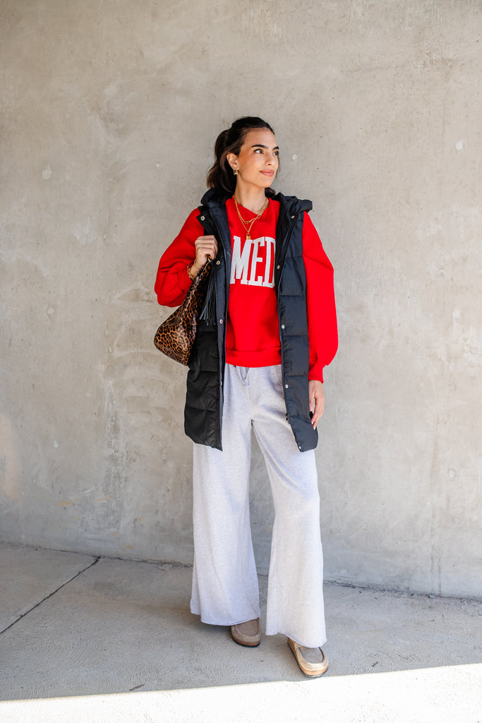 Woman wearing a red sweatshirt with text, black vest, white pants, and holding a brown bag against a plain wall.