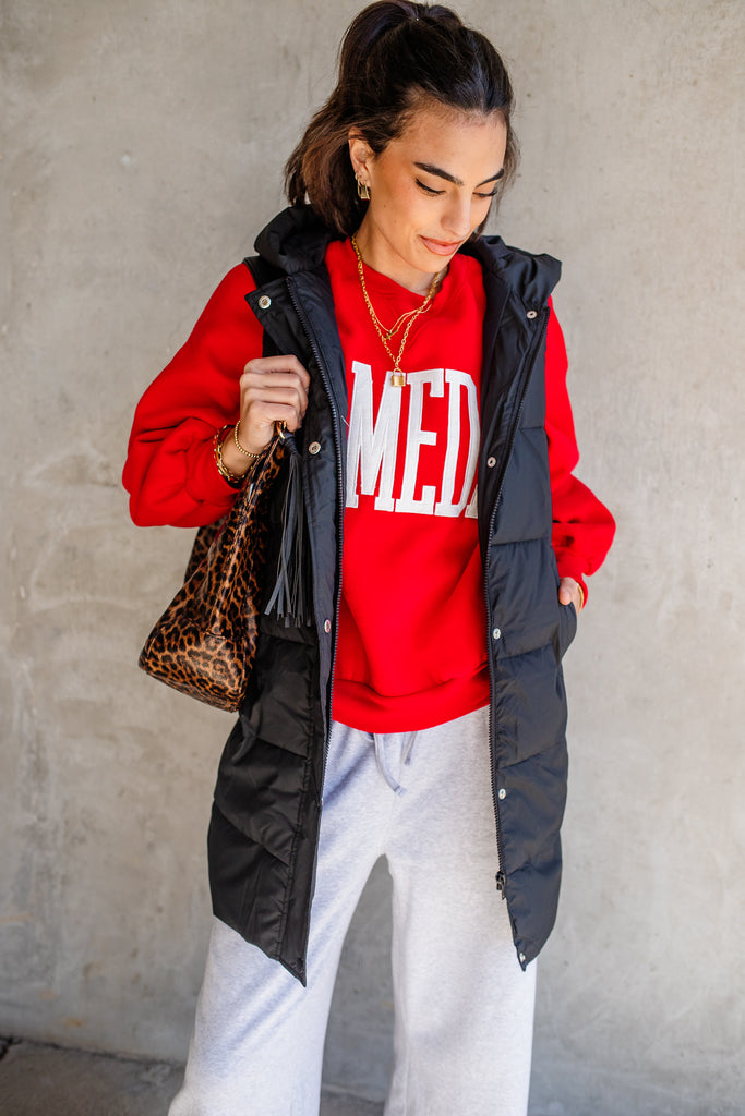 Woman wearing a red sweatshirt with, black vest, and white pants against a gray wall.