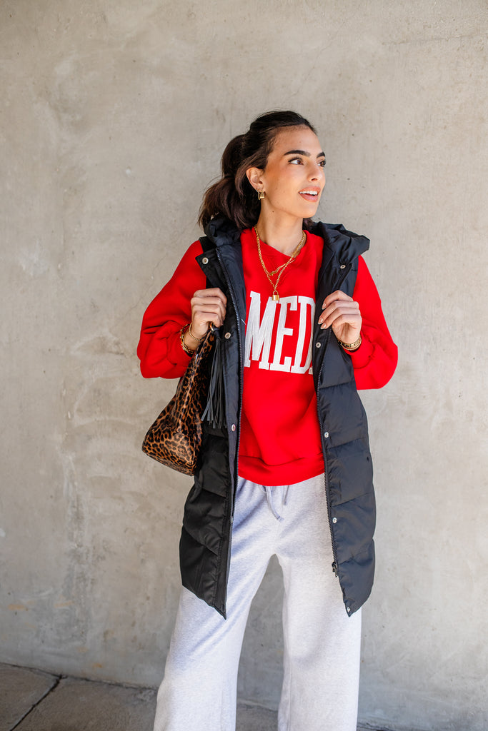 Woman wearing a red sweatshirt, black puffer vest, and white pants against a gray wall.