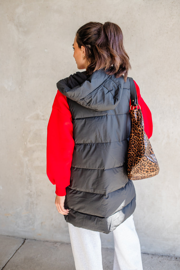 Person wearing a gray puffer vest over a red shirt with a leopard print bag, standing against a plain wall.