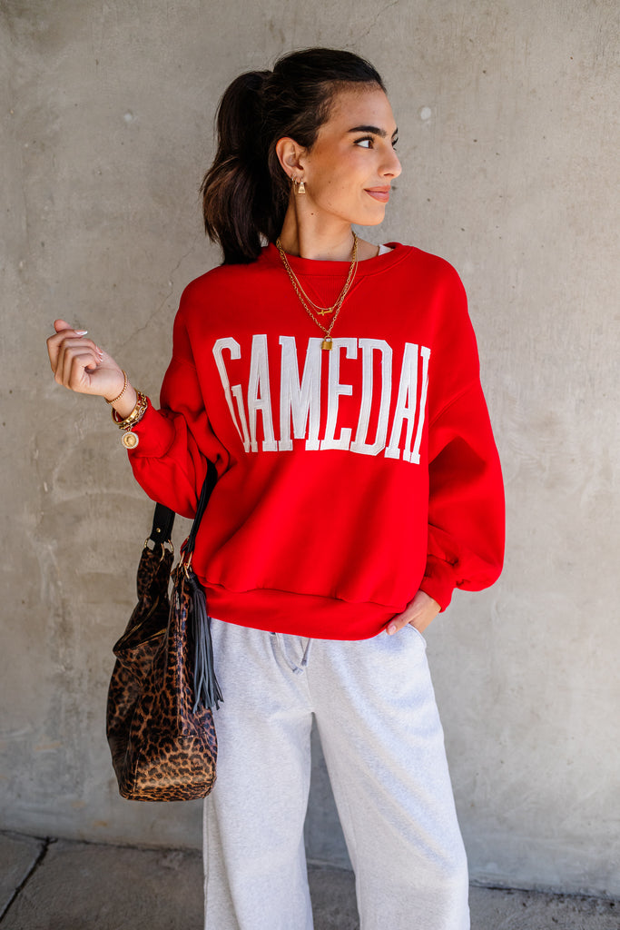 Woman wearing a red sweatshirt with 'GAMEDAY' text, standing against a concrete wall.