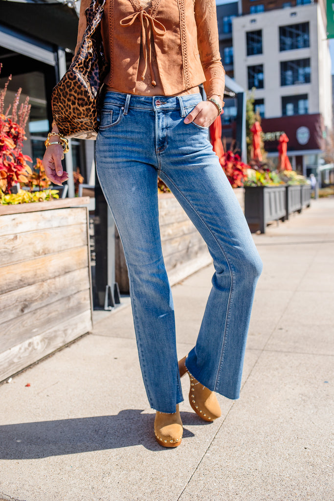 Person wearing a brown top, blue jeans, and tan shoes on a city street.