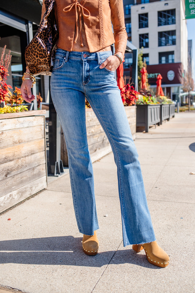 Person wearing a brown top and blue jeans standing on a sidewalk with buildings and plants in the background.