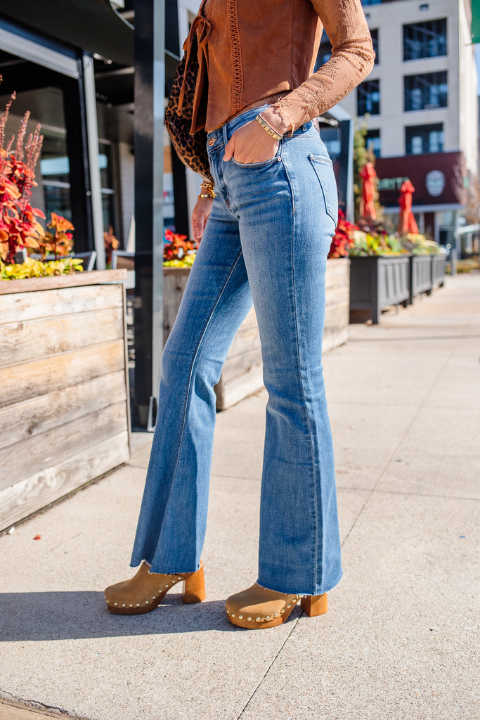 Person wearing a brown leather jacket, blue jeans, and brown shoes on a sidewalk.