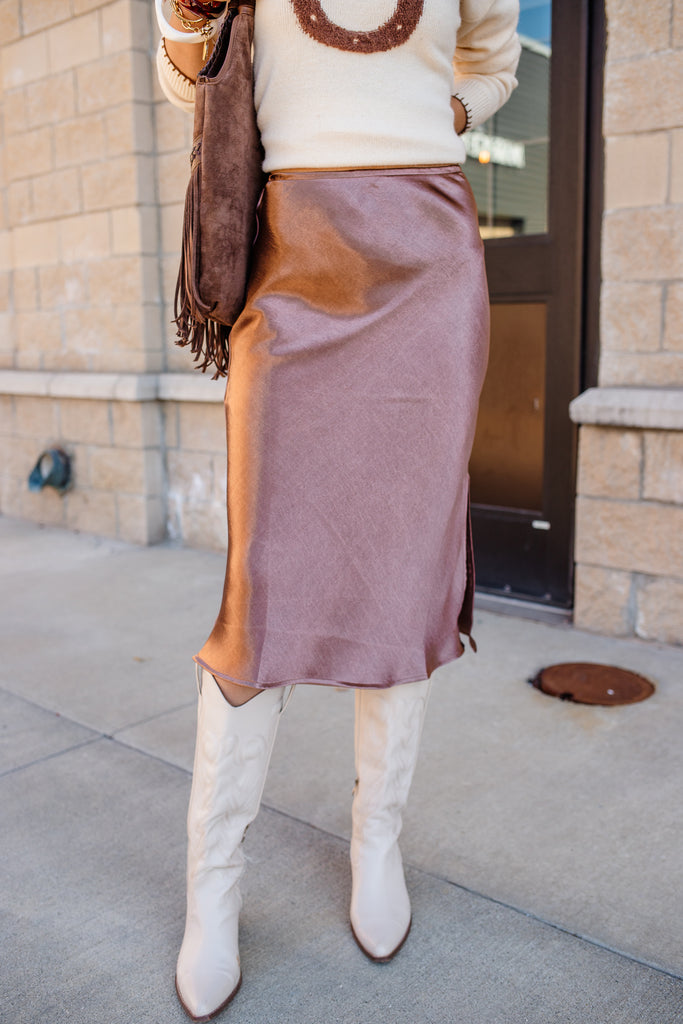 Person wearing a white sweater, brown skirt, and white knee-high boots standing outside a building.