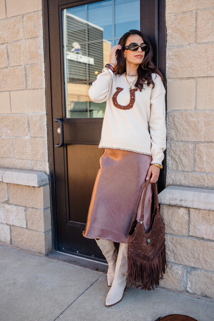Woman wearing a white sweater with a horse shoe design, brown leather skirt, and white boots, standing against a stone wall.