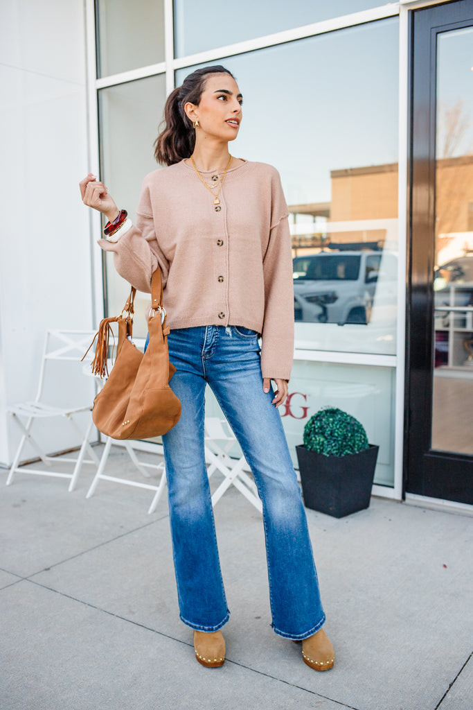 Woman wearing a beige cardigan, blue jeans, and brown shoes, holding a brown bag, standing outside a building.