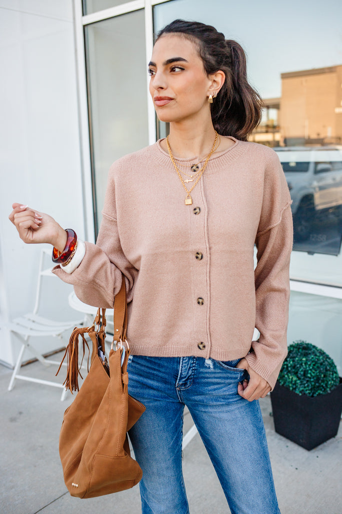 Woman wearing a beige cardigan and blue jeans, holding a brown handbag outdoors.