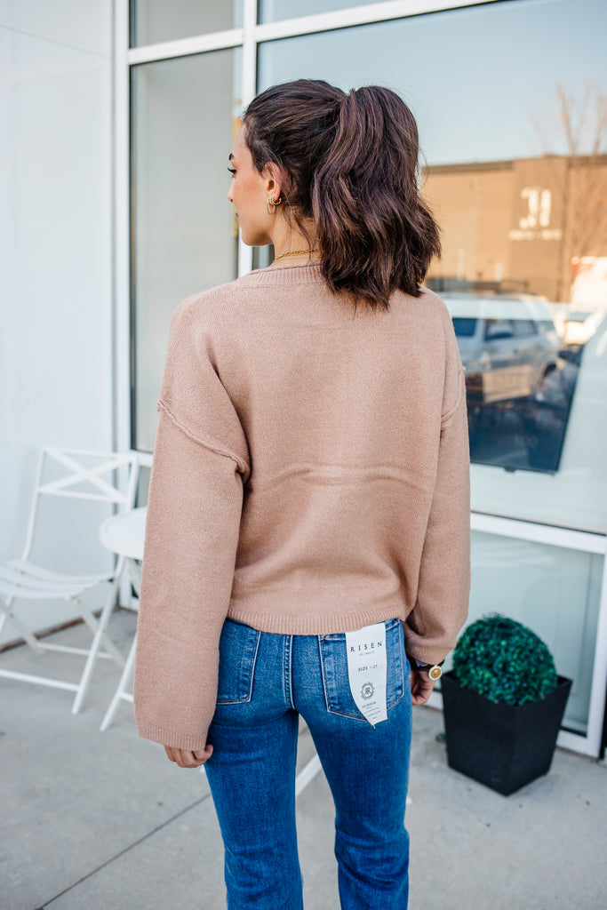 Woman wearing a beige sweater and blue jeans with a visible brand tag, standing outside a building.
