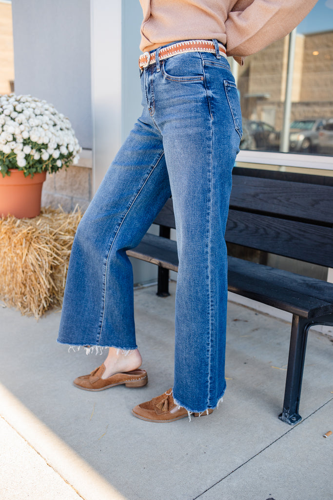 Person wearing blue jeans standing on a sidewalk with a bench and potted plant in the background.