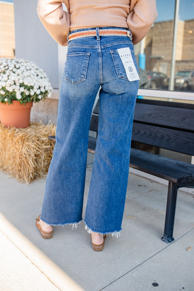 Person wearing blue jeans with a visible brand tag, standing outdoors near a bench and potted plant.