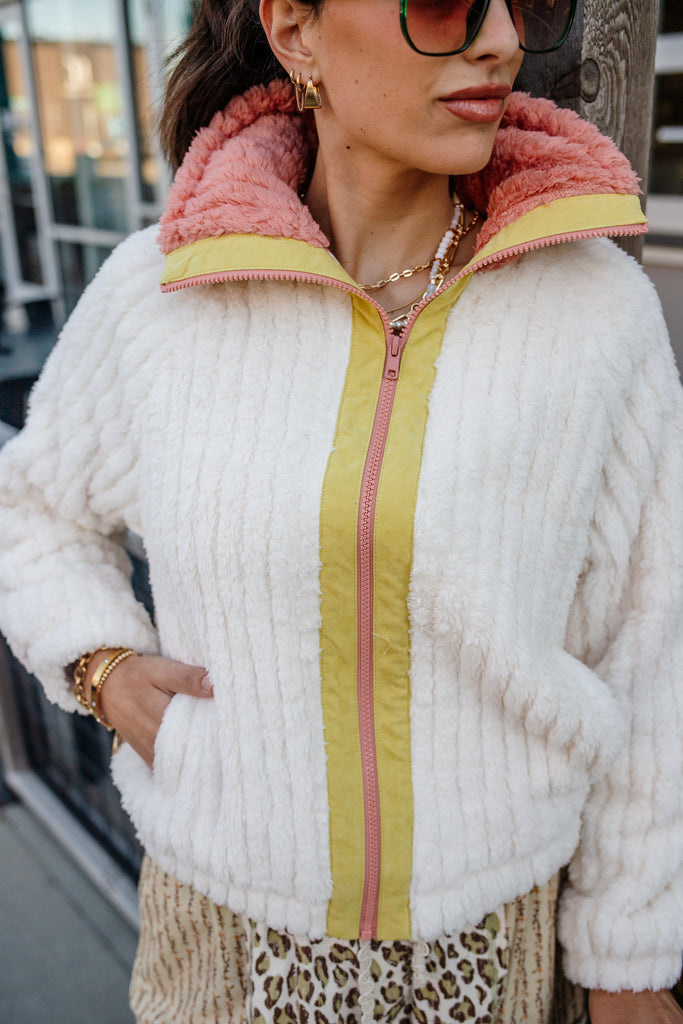 Woman wearing a white fluffy jacket with pink and yellow accents outdoors.