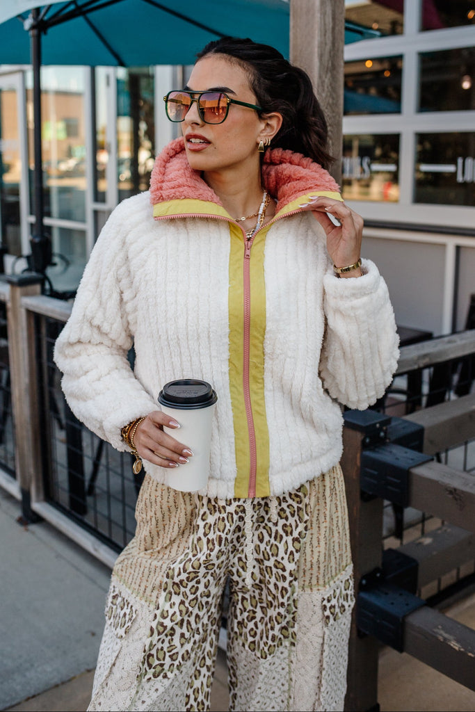 Woman in a stylish outfit with a white jacket, pink scarf, and leopard print pants holding a coffee cup outdoors.