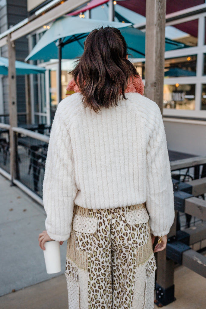Person wearing a white sweater and leopard print pants standing outdoors with tables and umbrellas in the background.